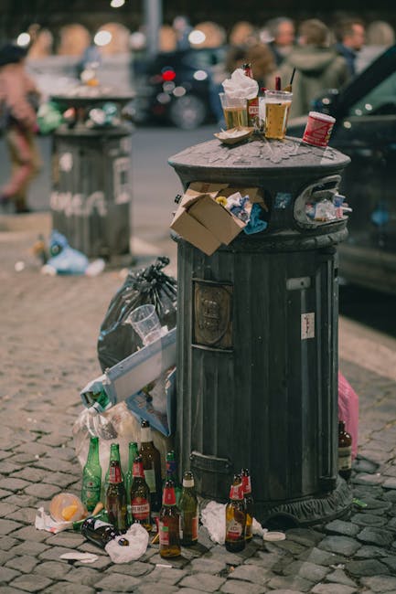 A city street scene showing a large, black, metal public rubbish bin that is overflowing with various waste items. On top of the bin, there are three disposable coffee cups with lids, two of which have plastic straws, and a crumpled paper napkin. Attached to the side of the bin are a few discarded cardboard food containers, with some spilling out onto the cobblestone pavement. Around the base of the bin, several empty glass bottles, including beer and soft drink bottles, are scattered, along with discarded paper towels and other small litter. In the background, there are a few parked cars, a pedestrian walking past, and a blurred view of other rubbish bins and urban elements, indicating an active city environment. The scene illustrates improper waste disposal practices, possibly related to take-away consumption, and highlights the presence of litter in public spaces, relevant to the scope of private rubbish collection and waste management services like those offered by Waste Collection Mayfair.