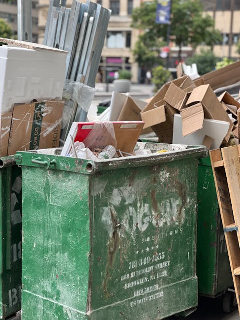 A large green waste collection bin situated on a city street, filled to the brim with various types of rubbish including flattened cardboard boxes, white styrofoam packaging, and loose paper. The bin's surface is weathered, with visible rust and dirt, indicating frequent use outside. Surrounding the bin are additional piles of discarded cardboard, some partially crushed or unfolded, and a few wooden pallets leaning against the bin on the right side. In the background, blurred urban elements such as trees, buildings, and parked cars can be seen, suggesting the scene is set in an urban environment where commercial or residential waste is being temporarily stored before collection. This scene exemplifies tangible aspects of private waste handling and on-site clearance, typical of rubbish removal services striving to prevent fly-tipping and improper disposal, with Waste Collection Mayfair providing professional solutions for managing such waste safely and efficiently.