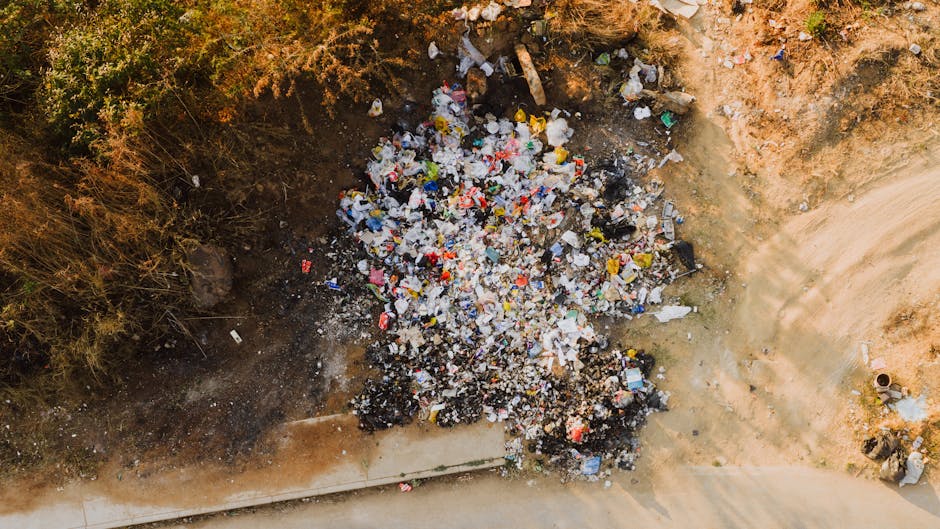 A city street scene showing a large, black, metal public rubbish bin that is overflowing with various waste items. On top of the bin, there are three disposable coffee cups with lids, two of which have plastic straws, and a crumpled paper napkin. Attached to the side of the bin are a few discarded cardboard food containers, with some spilling out onto the cobblestone pavement. Around the base of the bin, several empty glass bottles, including beer and soft drink bottles, are scattered, along with discarded paper towels and other small litter. In the background, there are a few parked cars, a pedestrian walking past, and a blurred view of other rubbish bins and urban elements, indicating an active city environment. The scene illustrates improper waste disposal practices, possibly related to take-away consumption, and highlights the presence of litter in public spaces, relevant to the scope of private rubbish collection and waste management services like those offered by Waste Collection Mayfair.