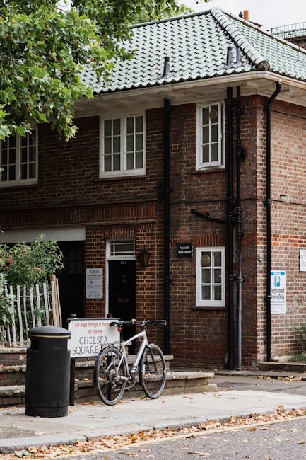 The image shows the exterior of a two-storey brick building with a grey tiled pitched roof accented by several small black vents. The building's dark-framed white sash windows are evenly spaced and located on both floors. To the right of the main door, which is set within a brick wall, there is a small white sign and a dark rectangular mailbox. Leaning against a black metal railing in front of the building is a white bicycle with black tires and a black saddle, positioned next to a black cylindrical trash bin. Nearby, a temporary white sign with red and black lettering indicates waste-related information, with the words 'Chelsea Square' partially visible. The scene includes a paved sidewalk with fallen autumn leaves scattered across it and a nearby fence with green foliage. The overall setting suggests an urban residential area where waste collection may be arranged privately, supported by the presence of waste management signage, and aligns with the context of on-site rubbish removal services facilitated by waste collection companies like Waste Collection Mayfair.