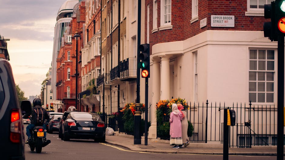 A street scene in a residential area showing a corner building with white and red brick facades, tall windows, and black wrought iron railings on the balconies. Two pedestrian traffic lights are visible, one displaying green and the other red. In the foreground, a woman wearing a pink coat and a headscarf stands on the sidewalk, next to a flower bed with orange and yellow flowers surrounded by a black metal fence. Several parked cars line the street, including a black sedan and a motorbike with a rider wearing a black helmet. The sky above is overcast with a warm, muted light, and a street sign indicating 'Upper Brook Street W1' is attached to the building's corner, suggesting an area in central London. The scene captures a typical urban environment suited to private waste collection and rubbish removal services, with a focus on pedestrian and vehicular activity in a historic residential neighborhood.