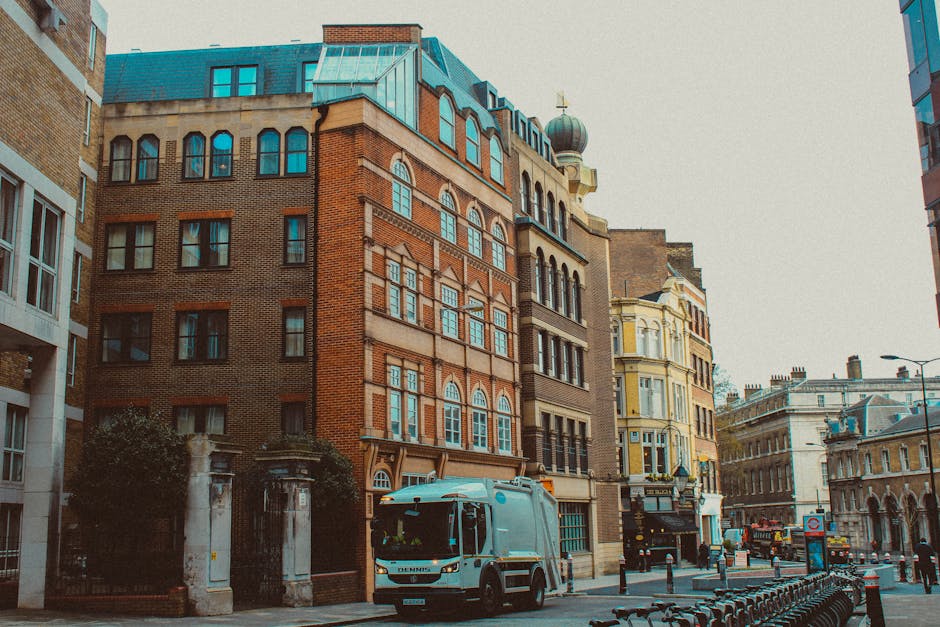 A small waste collection van with a light blue body and white roof parked on a city street in front of multi-story buildings with various architectural styles, including brick and stucco facades. The vehicle is positioned near a sidewalk with a row of black metal bollards, and behind it, there are additional buildings featuring large windows with arches and decorative elements. On the left, part of a gated entrance is visible, with stone pillars and greenery nearby. The street scene includes a few bicycles parked along the curb, and an urban environment characterized by a mix of historic and modern structures under overcast sky lighting. The scene suggests an area where private waste collection services might operate as part of a broader urban waste management approach.