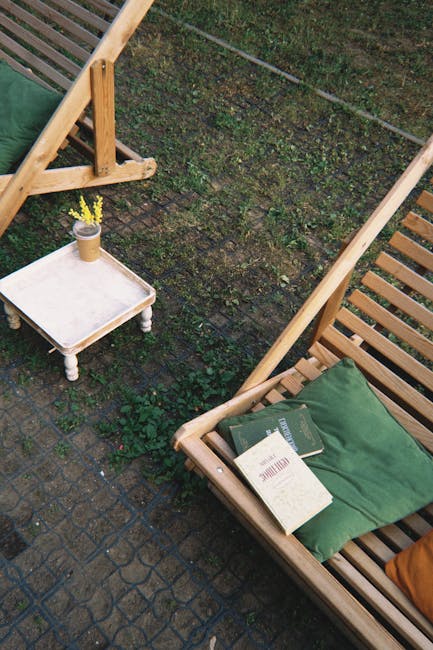 The image depicts an outdoor patio or garden area with two wooden lounge chairs featuring slatted designs and green cushions placed on their seats. The chairs are positioned on a paved surface made of dark, textured bricks. A small, white, rectangular side table with turned legs is situated nearby, holding a yellow flower in a small pot. The background shows a grassy border with patches of moss and small plants, bordered by a stone or concrete edge, and a thin wire or string runs diagonally across the scene. The overall setting appears to be a private outdoor space, with no visible waste or rubbish, but the arrangement and context subtly suggest a location where private waste handling or on-site disposal could be relevant, aligning with the themes of rubbish removal services. The lighting is natural, indicating a cloudy or overcast day, creating a neutral and calm atmosphere. Waste Collection Mayfair’s service context is reflected through the outdoor environment, where furniture and minimal clutter suggest tidiness and potential preparation for waste collection or disposal activities related to outdoor furniture or garden maintenance.