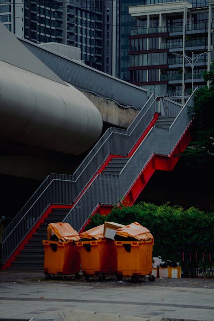 In the foreground of the image, there are three large, bright orange waste bins made of durable plastic, positioned on the paved ground and filled with various types of rubbish, indicative of a waste collection or disposal area outside an urban building. Behind the bins, a green shrub provides some natural contrast to the industrial scene. The central focus is on a modern external staircase with metal handrails and steps, painted in gray with a striking red underside that leads upwards alongside a large cylindrical pipeline or duct running horizontally above it. The staircase is attached to a multi-story building in the background, characterized by dark glass windows and a contemporary architectural design with multiple balconies and structural beams. The overall setting appears to be an urban environment, likely a commercial or residential complex, with controlled environmental lighting suggesting daylight, and the scene emphasizing the practical aspect of waste management in a city setting, featuring elements of independent waste collection outside standard municipal services.
