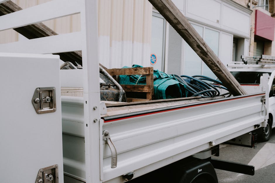 The image depicts the side of a white flatbed utility vehicle parked along a city street, loaded with various items for waste removal. Visible on the flatbed are a weathered wooden pallet, coiled black hoses, a folded green tarp or fabric, and a partly visible metallic tool or container, all arranged in a somewhat haphazard manner. The surrounding environment includes a modern building facade with large windows, some with unconventional glass panels and external shading elements, hinting at an urban or residential setting. The vehicle's side panel features several metal fasteners and a handle, and the flatbed appears to be designed for carrying bulky waste or construction debris, indicative of private or independent rubbish collection services. The lighting is natural, with a neutral tone, emphasizing the textures of the materials: the rough finish of the wooden pallet, the flexible coating of the hoses, and the smooth paint of the vehicle. This scene represents an organized approach to on-site clearance and waste handling, typical of services like those offered by Waste Collection Mayfair for bulky or debris removal.