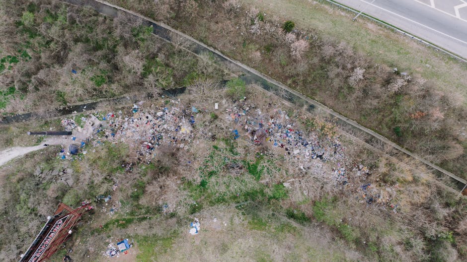 An aerial view of a large, unmanaged accumulation of mixed household and commercial waste scattered across a patch of land adjacent to a paved pathway and a railway track. The rubbish includes numerous plastic bags, containers, cardboard boxes, and miscellaneous debris, creating a dense, cluttered area with varying heights of accumulated waste. The environment features leafless trees and sparse greenery, with some small patches of grass and bushes amidst the litter. To the upper right, a road runs parallel to the railway line, separated by a narrow strip of grass and a fence. The overall scene highlights improper waste disposal outside authorized collection zones, emphasizing the need for professional rubbish removal services such as those offered by Waste Collection Mayfair, which specialize in private waste management solutions to prevent illegal dumping and maintain environmental standards.