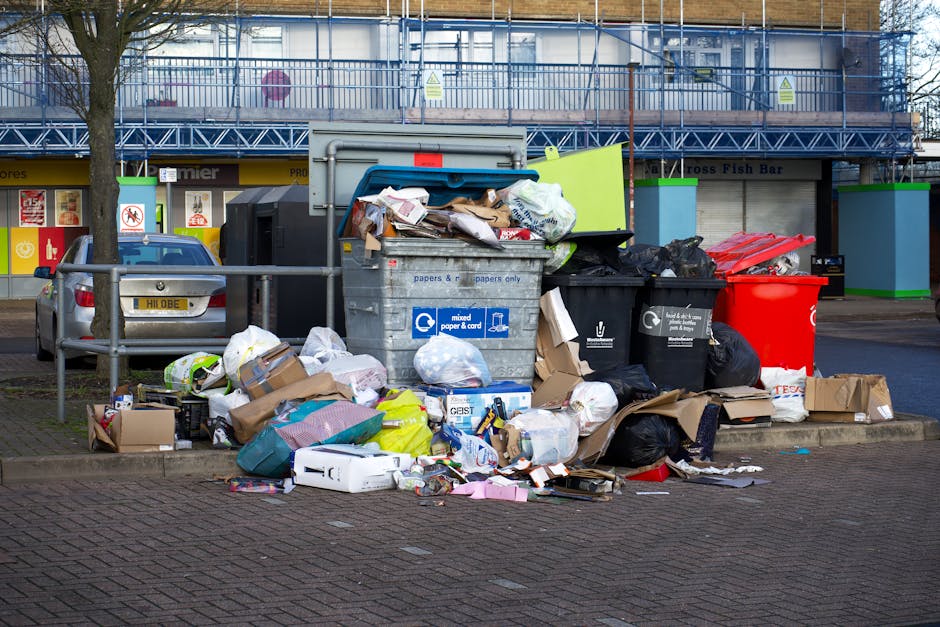 A collection of overflowing rubbish bins and scattered waste on a paved urban street, with various types of trash including cardboard boxes, plastic bags, paper waste, and discarded packaging predominantly in brown, black, and white colors. The large grey bin labeled for mixed paper and card is positioned at the center, with its lid open and paper scraps visible spilling onto the ground. Adjacent to it are black recycling bins and a bright red bin, all filled and overflowing with rubbish. Surrounding the bins, loose waste and packaging materials are strewn across the pavement, contributing to a cluttered scene. In the background, a building with a shopfront featuring signage, a parked silver vehicle, and a metal railing separates the rubbish area from the street, with a tree trunk visible on the left side. The environment appears to be in daytime with natural light, reflecting an aspect of private waste collection typical of independent rubbish removal services such as those provided by Waste Collection Mayfair.