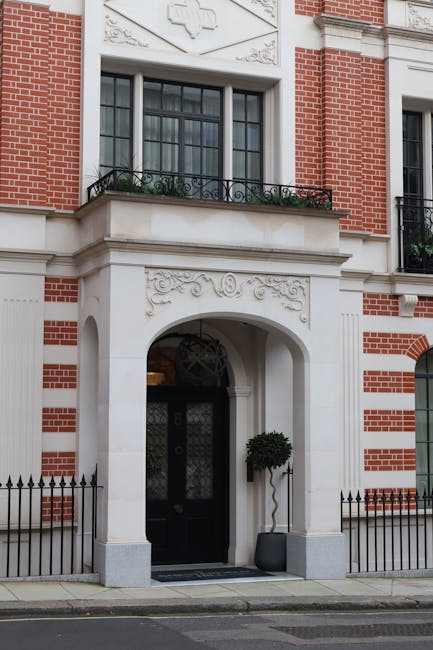 The image depicts the façade of a classic-style building with a red brick exterior complemented by white decorative stonework and mouldings. The main entrance features a large, arched doorway painted in dark color, with a decorative transom window above, framed by ornate white carvings. To the left of the door, a small, round, well-maintained shrub is placed inside a modern, dark, rectangular planter. Above the entrance, a balcony with black wrought iron railing extends along the façade, supporting a large window with multiple glass panes divided by black muntins, set within a white frame. The building’s exterior wall includes detailed carved motifs, and the window has dark glass panes with a faint reflective quality. Natural daylight illuminates the scene, revealing the building’s clean and well-maintained condition. The ground in front of the entrance is paved with concrete, and a low black metal fence runs along the sidewalk, providing a boundary for the property. The overall setting suggests an urban residential or commercial property that may require occasional waste removal or on-site clearance, consistent with services provided by Waste Collection Mayfair.