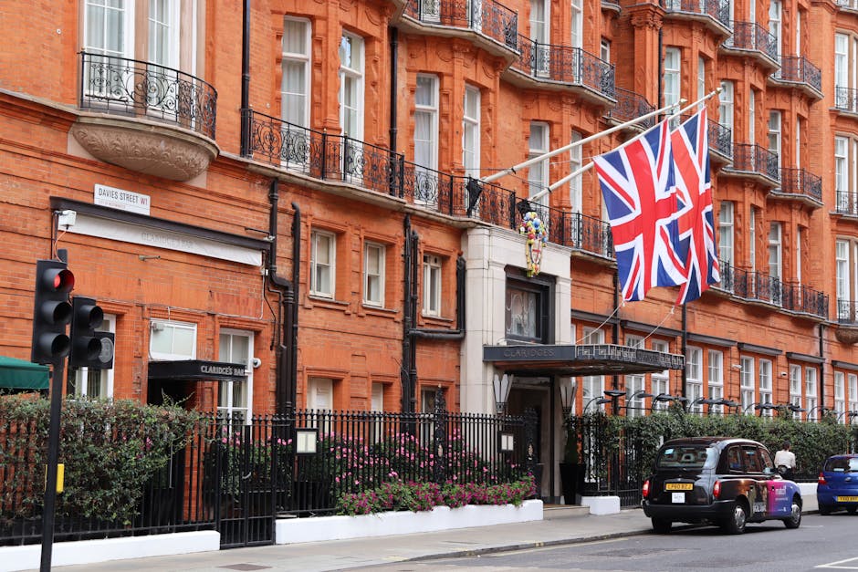 The image depicts the exterior of a red-brick multi-storey building with ornate black iron railings on the balconies and white-framed sash windows. Two British flags are prominently displayed on a flagpole extending from the building facade, fluttering against a partly cloudy sky. In the foreground, a black fence with pointed finials surrounds a small garden area densely planted with bushes and flowering plants, which appear to be pink and purple skimmia or similar shrubbery. To the left, a traffic light is visible, showing a red signal, situated next to a street sign indicating 'Davies Street W1'. The building's entrance features a covered porch with a black canopy displaying the name 'Claridge's' in silver lettering, suggesting a hotel or prestigious establishment. Parked along the street are a dark-colored taxi and a few other vehicles, including a blue car further down, with pedestrians walking past. The scene captures an urban environment with a combination of residential and commercial characteristics, aligned with private waste collection and rubbish removal services operating in central London areas.