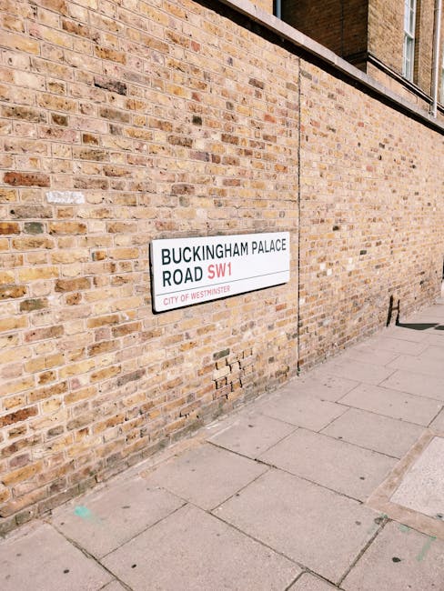 A brick wall on a sidewalk features a white street sign with black and red text indicating 'BUCKINGHAM PALACE ROAD SW1' in Westminster, suggesting the location is near Buckingham Palace. The wall's bricks are a mix of light brown, beige, and reddish hues with a rough texture, some bricks showing minor chips and wear. The sign is mounted flush against the wall, positioned slightly above the sidewalk, which is paved with large concrete slabs that show slight discoloration and minor cracks. The pavement extends to the right, with a narrow strip of curb visible along the edge. In the background, part of a building with a window is visible, indicating an urban environment. The scene is lit by natural daylight, casting subtle shadows on the wall and pavement, and it provides context for urban waste management or rubbish removal services, such as those offered by Waste Collection Mayfair, operating in the area.